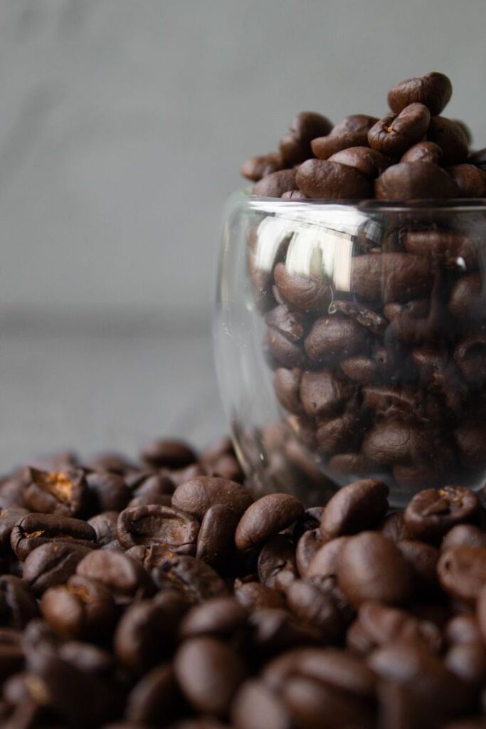 Glass cup full of roasted brown coffee beans placed on table against gray background in daylight